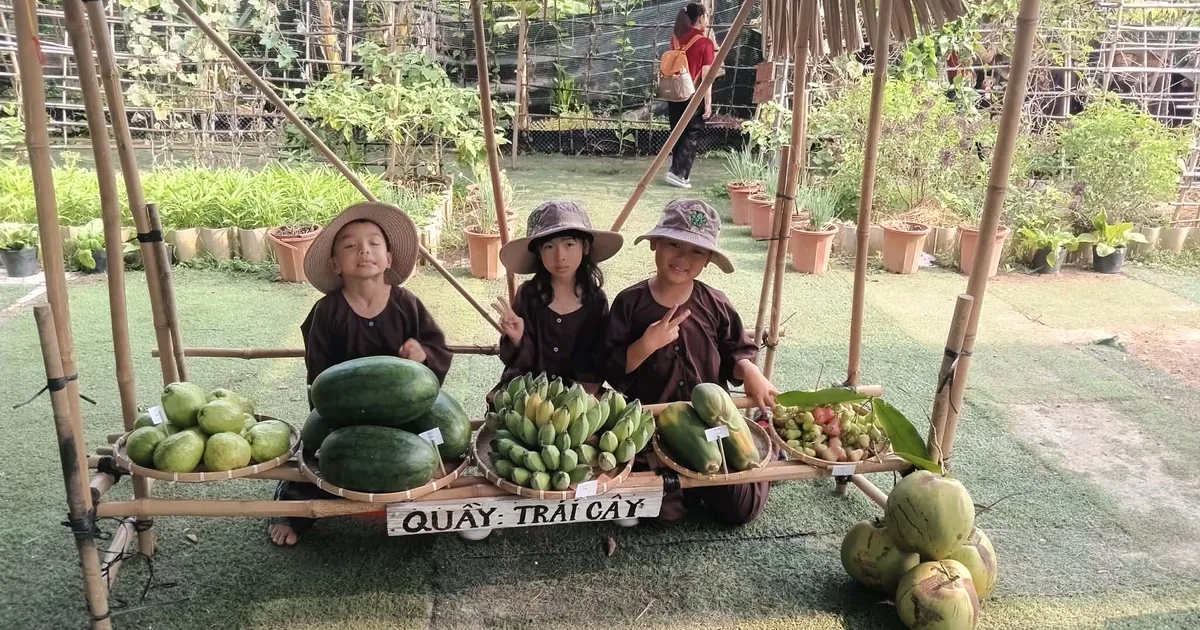 Ba bé mặc áo bà ba ngồi sau quầy trái cây tại chợ quê Dragon Farm